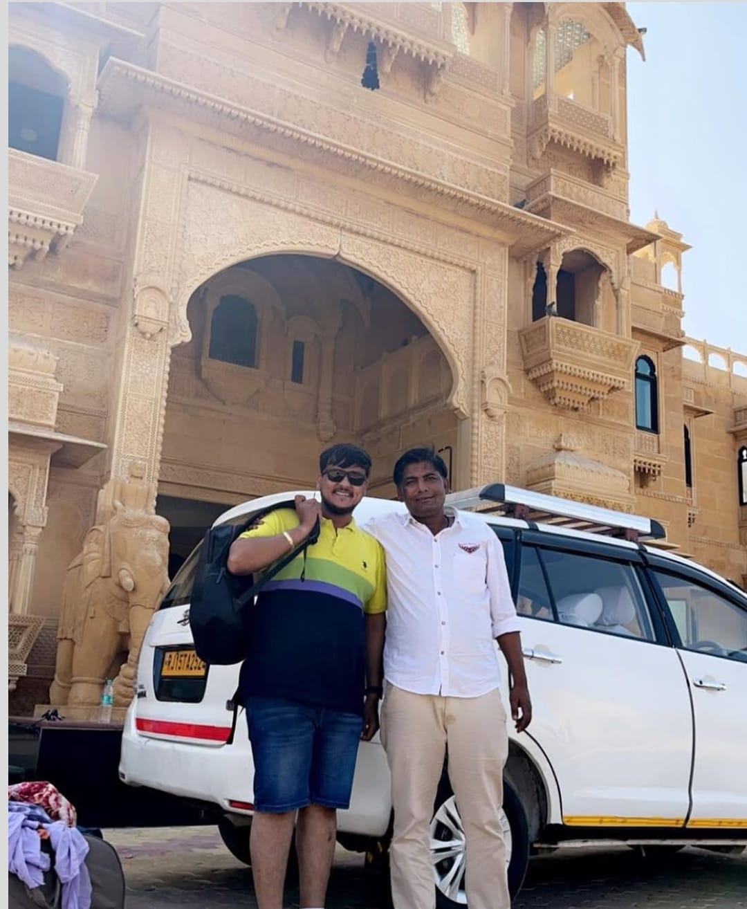 Two people standing beside a white travel vehicle in front of ornate sandstone architecture.