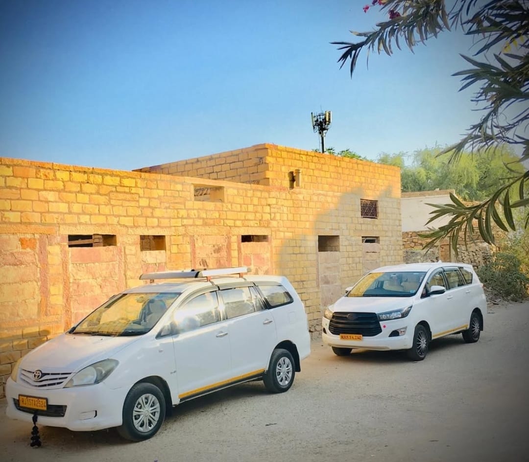 Two white travel vehicles parked beside a sandstone building in the Jaisalmer region.